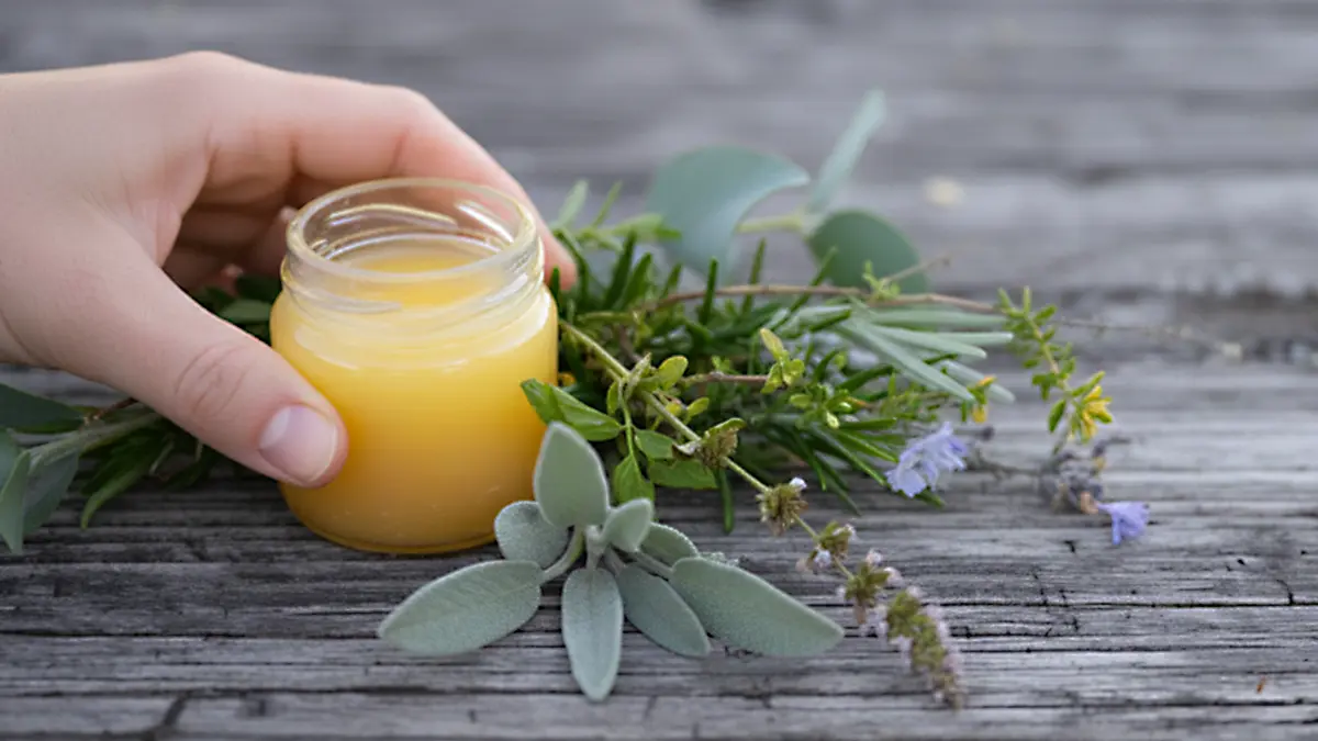 Hand hält selbst gemachten gelben Kräuterbalsam im Glas, daneben frische Winterkräuter auf Holzuntergrund