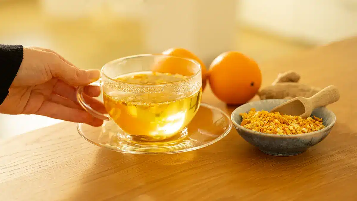 Warmer Orangenschalen-Tee in einer Glastasse auf einem Holztisch, mit frischen Orangen und getrockneten Orangenschalen